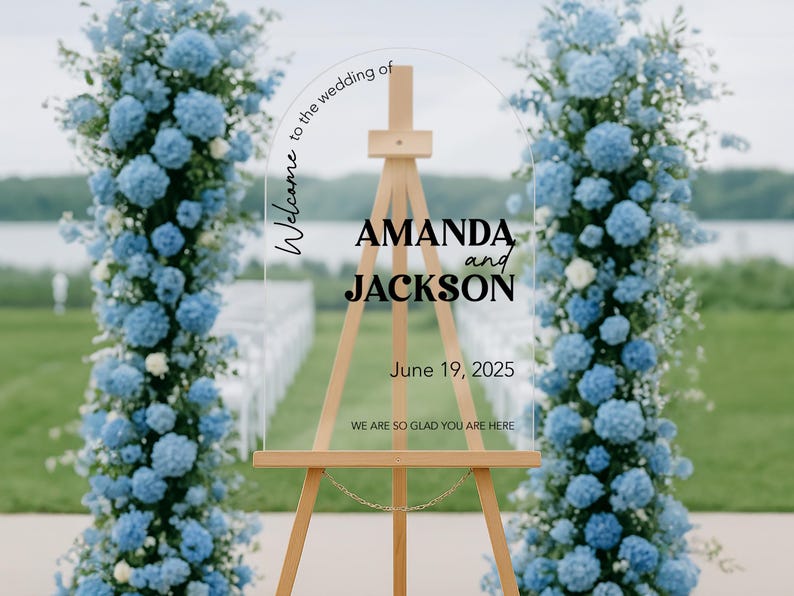 May include: A clear acrylic wedding sign on a wooden easel, with the names "AMANDA and JACKSON" and the date "June 19, 2025". The sign is surrounded by blue floral arrangements. Text reads "Welcome to the wedding of" and "WE ARE SO GLAD YOU ARE HERE".