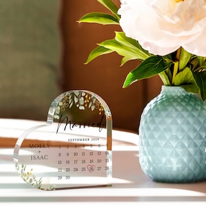 Puede incluir: Un recuerdo de boda de acr&iacute;lico transparente en forma de coraz&oacute;n con un dise&ntilde;o floral y las palabras "Casado" y "Septiembre 2024". El calendario muestra la fecha "26" rodeada de un c&iacute;rculo rojo. Los nombres "Molly + Isaac" est&aacute;n impresos debajo del calendario.