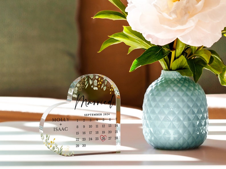 Puede incluir: Un recuerdo de boda de acr&iacute;lico transparente en forma de coraz&oacute;n con un dise&ntilde;o floral y las palabras "Casado" y "Septiembre 2024". El calendario muestra la fecha "26" rodeada de un c&iacute;rculo rojo. Los nombres "Molly + Isaac" est&aacute;n impresos debajo del calendario.