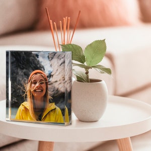 May include: A clear, square crystal photo block displaying a smiling person in a yellow jacket and orange beanie. The photo block sits on a white side table next to a small potted plant and a reed diffuser.