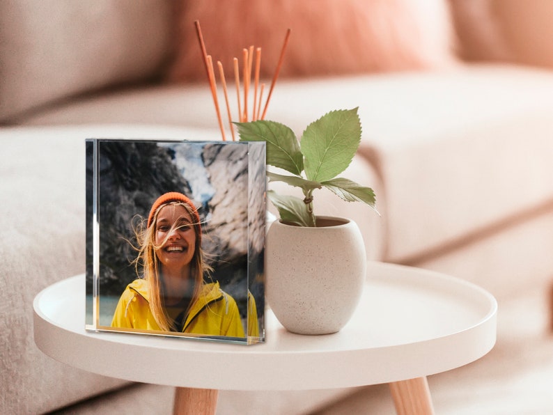 May include: A clear, square crystal photo block displaying a smiling person in a yellow jacket and orange beanie. The photo block sits on a white side table next to a small potted plant and a reed diffuser.