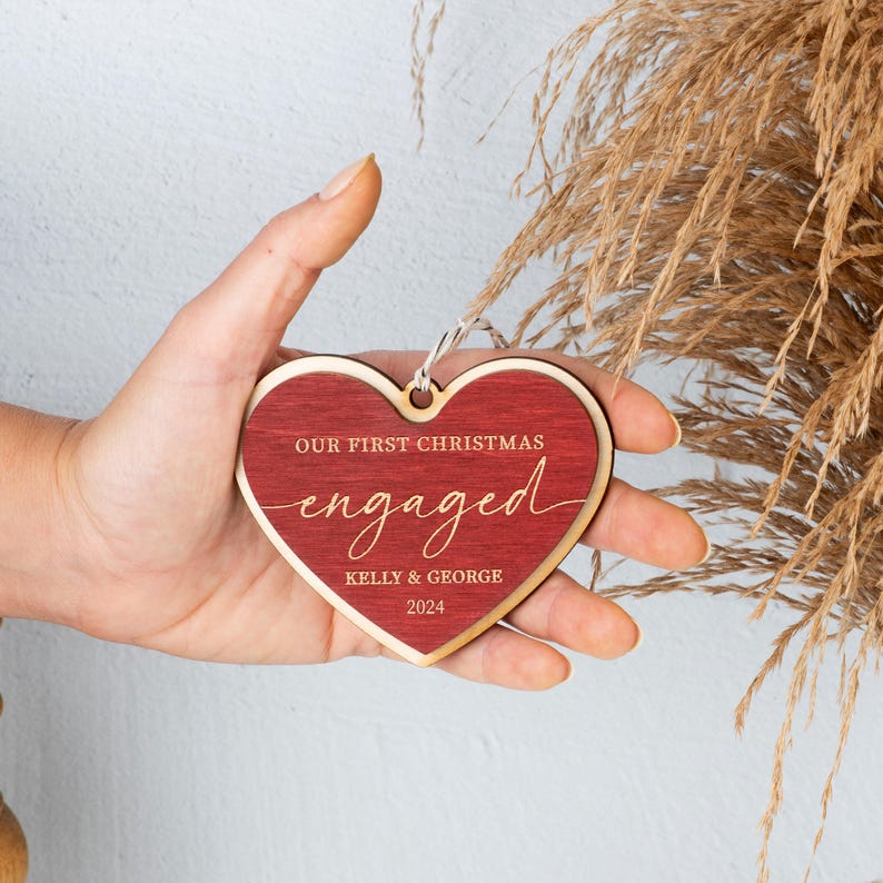 May include: Red wooden heart-shaped ornament with the text "Our First Christmas Engaged Kelly & George 2024" in gold lettering.