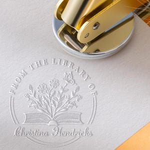 May include: A gold-colored embosser rests on a white paper with a floral and butterfly design. The embossed text reads "FROM THE LIBRARY OF" above an open book with flowers and a butterfly, and the name "Christina Hendricks" below.