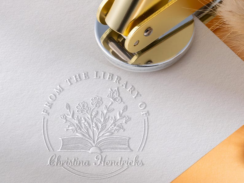 May include: A gold-colored embosser rests on a white paper with a floral and butterfly design. The embossed text reads "FROM THE LIBRARY OF" above an open book with flowers and a butterfly, and the name "Christina Hendricks" below.