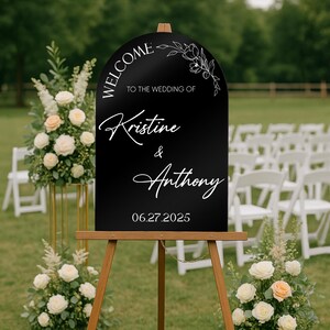 May include: A black wedding welcome sign on a wooden easel. The sign has white script that reads "Welcome to the wedding of Kristine & Anthony 06.27.2025". Floral arrangements and white chairs are in the background.