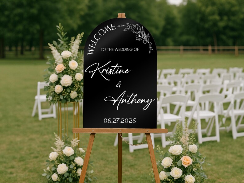 May include: A black wedding welcome sign on a wooden easel. The sign has white script that reads "Welcome to the wedding of Kristine & Anthony 06.27.2025". Floral arrangements and white chairs are in the background.