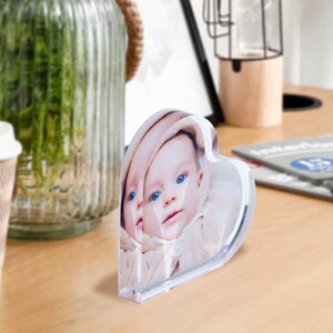 May include: A heart-shaped acrylic photo block featuring a baby's face with blue eyes. The clear block stands upright on a wooden surface, reflecting the image. A coffee cup and a glass vase are in the background.
