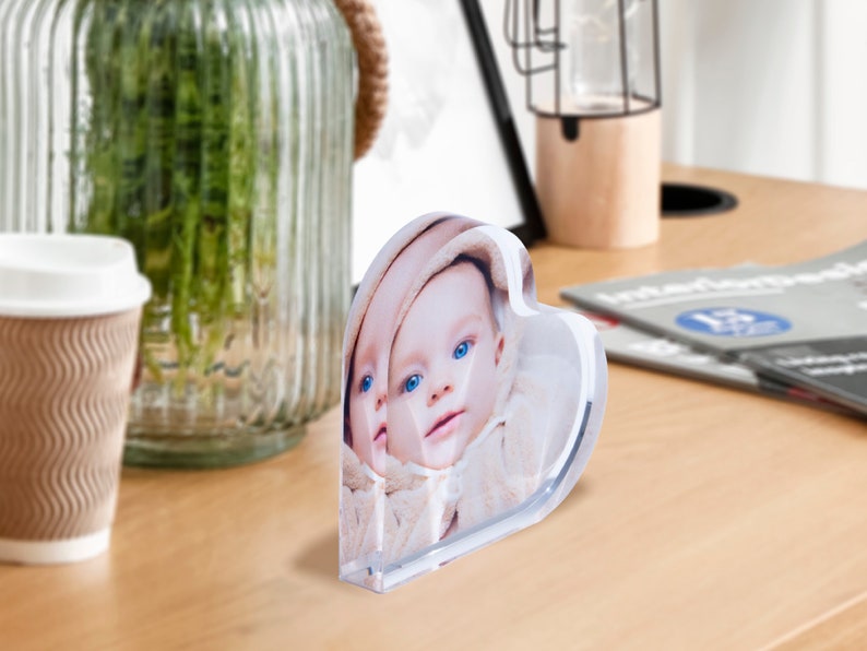 May include: A heart-shaped acrylic photo block featuring a baby's face with blue eyes. The clear block stands upright on a wooden surface, reflecting the image. A coffee cup and a glass vase are in the background.