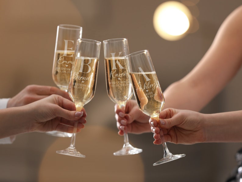 Może przedstawiać: Cztery kieliszki do szampana wzniesione w geście toastu. Każdy kieliszek jest grawerowany imieniem, tytułem i datą. Kieliszki są wypełnione musującym napojem. Tło jest rozmyte, z miękkim oświetleniem.