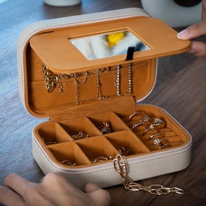 K&ouml;nnte beinhalten: Eine wei&szlig;-beige Schmuckschatulle mit offenem Deckel, die einen Spiegel und F&auml;cher freigibt. Der Innenraum ist beige und die Schatulle enth&auml;lt verschiedene goldene Halsketten, Armb&auml;nder und Ringe. Die Schatulle steht auf einer Holzoberfl&auml;che.