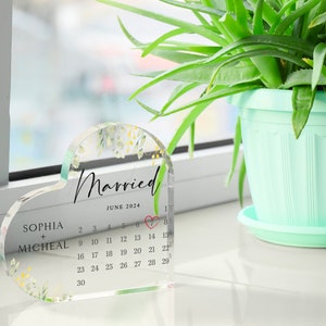Puede incluir: Un recuerdo de boda de acr&iacute;lico transparente en forma de coraz&oacute;n con un dise&ntilde;o floral y un calendario para junio de 2024. El texto "Married" est&aacute; escrito en cursiva en la parte superior del calendario. Los nombres "Sophia + Micheal" est&aacute;n escritos debajo del calendario.