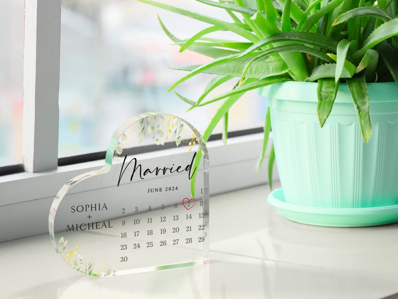 Puede incluir: Un recuerdo de boda de acr&iacute;lico transparente en forma de coraz&oacute;n con un dise&ntilde;o floral y un calendario para junio de 2024. El texto "Married" est&aacute; escrito en cursiva en la parte superior del calendario. Los nombres "Sophia + Micheal" est&aacute;n escritos debajo del calendario.