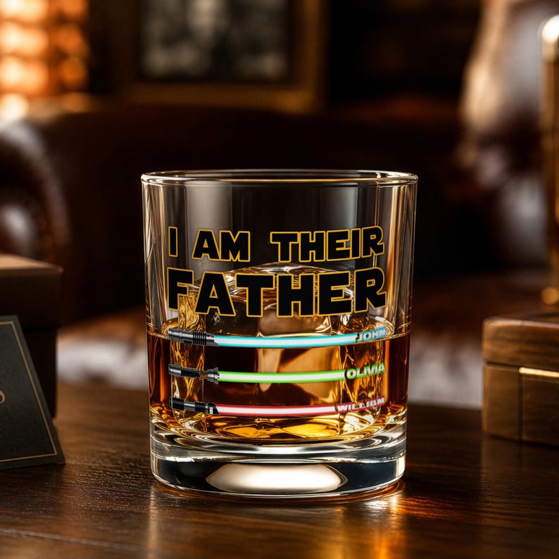 May include: A clear rocks glass with the text "I AM THEIR FATHER" in black and gold. The glass features three lightsaber designs in blue, green, and red, with names below. The glass is filled with a brown liquid.