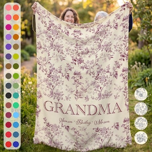 May include: A cream-colored blanket with a floral pattern in shades of purple. The word "GRANDMA" is printed in large letters, with names below. The blanket is held up by two people outdoors.