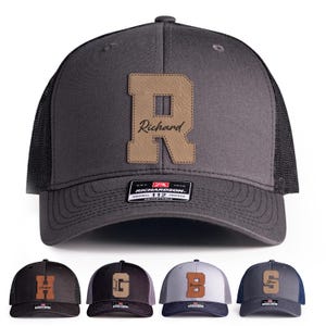 May include: Gray trucker hat with a leather patch featuring the letter "R" and the name "Richard". The hat has a black mesh back and an adjustable strap. Additional hats with different letters and names are displayed below.