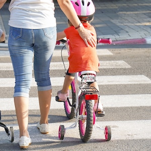 May include: A young child wearing a pink helmet rides a white bicycle with training wheels. The child is wearing an orange shirt and is being helped by an adult wearing blue jeans.