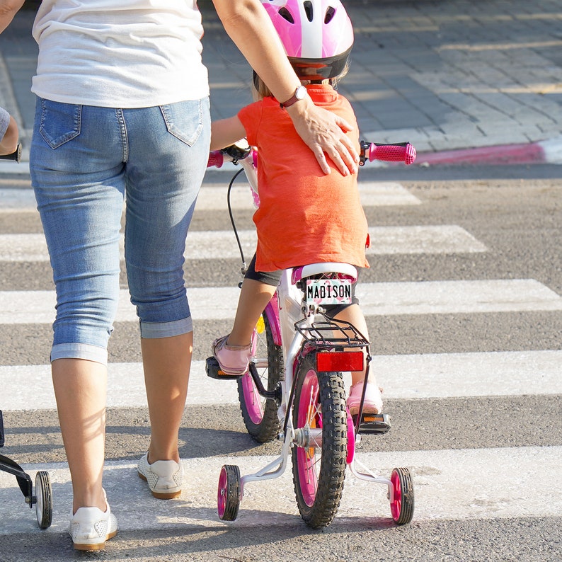 May include: A young child wearing a pink helmet rides a white bicycle with training wheels. The child is wearing an orange shirt and is being helped by an adult wearing blue jeans.