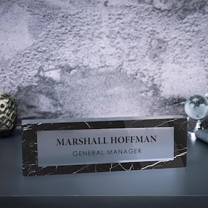 May include: Black and white marble desk nameplate with a clear acrylic insert. The text on the nameplate reads "Marshall Hoffman, General Manager".