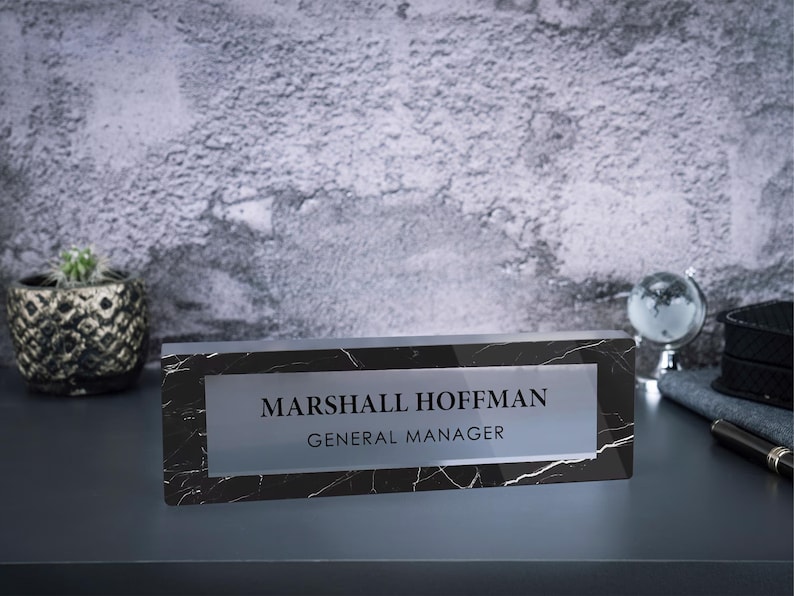 May include: Black and white marble desk nameplate with a clear acrylic insert. The text on the nameplate reads "Marshall Hoffman, General Manager".