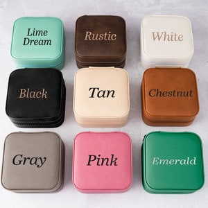 K&ouml;nnte beinhalten: Eine Sammlung von neun quadratischen Schmuckk&auml;stchen in verschiedenen Farben, darunter Limettengr&uuml;n, Braun, Wei&szlig;, Schwarz, Beige, Kastanienbraun, Grau, Rosa und Smaragdgr&uuml;n. Jedes K&auml;stchen hat einen Rei&szlig;verschluss und einen farblich passenden gestickten Namen.