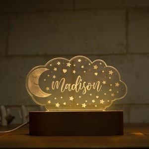 May include: A night light shaped like a cloud with a crescent moon and stars. The name "Madison" is written in script across the center. The light sits on a rectangular wooden base and is plugged in.