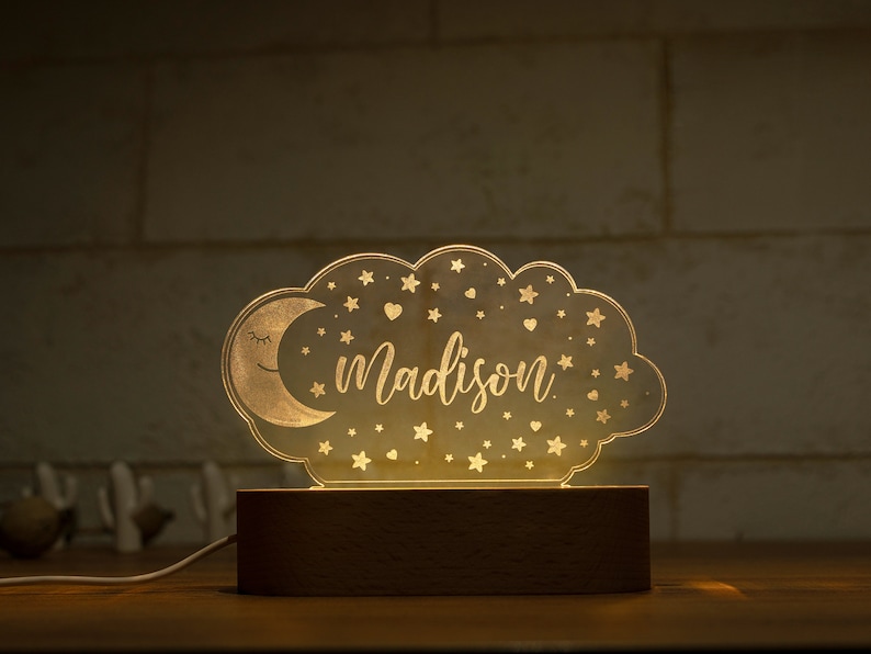 May include: A night light shaped like a cloud with a crescent moon and stars. The name "Madison" is written in script across the center. The light sits on a rectangular wooden base and is plugged in.