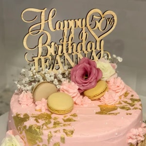 May include: A pink frosted cake decorated with gold leaf, macarons, and fresh flowers. A wooden cake topper reads "Happy 50th Birthday JEANNA" in elegant script. The cake is on a white cake stand.