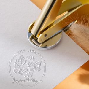 May include: A gold-colored library seal is pressed onto a white paper, creating an embossed design. The design includes three butterflies, a leafy branch, and the text "FROM THE LIBRARY OF Jessica Williams."