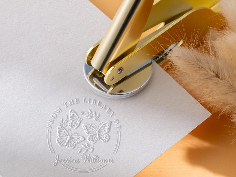 May include: A gold-colored library seal is pressed onto a white paper, creating an embossed design. The design includes three butterflies, a leafy branch, and the text "FROM THE LIBRARY OF Jessica Williams."
