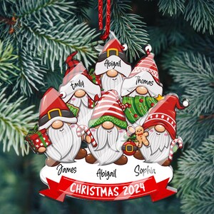 May include: A Christmas ornament featuring a group of six gnomes with red hats and white beards. Each gnome has a name written on their hat: Abigail, Emily, Thomas, James, Abigail, and Sophia. The ornament also includes the text "CHRISTMAS 2024".
