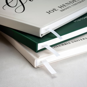 May include: Three hardcover books stacked on top of each other. The top book is beige with the text "Joe Hende Brown University" printed on the cover. The middle book is dark green with the text "Midtown High School Class of 20" printed on the cover. The bottom book is beige with the text "Columbia University Class" printed on the cover.