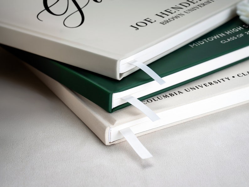 May include: Three hardcover books stacked on top of each other. The top book is beige with the text "Joe Hende Brown University" printed on the cover. The middle book is dark green with the text "Midtown High School Class of 20" printed on the cover. The bottom book is beige with the text "Columbia University Class" printed on the cover.