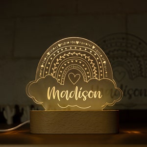 May include: A decorative rainbow-shaped night light with the name "Madison" illuminated in a warm glow. The design features a rainbow with heart and leaf details, set on a wooden base. The light is connected to a white cord.