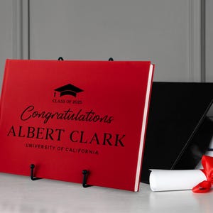 May include: Red graduation guest book with black text that reads "Congratulations Albert Clark, University of California, Class of 2023".