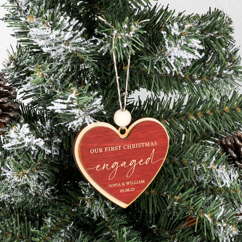 May include: Red wooden heart-shaped Christmas ornament with the text "Our First Christmas engaged" and the names "Sofia & William" and the date "05.08.23".
