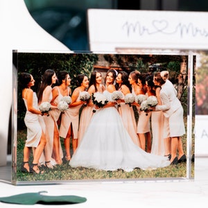 May include: A clear acrylic photo block displaying a wedding photo. The image features a bride in a white gown surrounded by bridesmaids in peach dresses, holding bouquets. A white vase and a framed sign with "Mr & Mrs" are in the background.