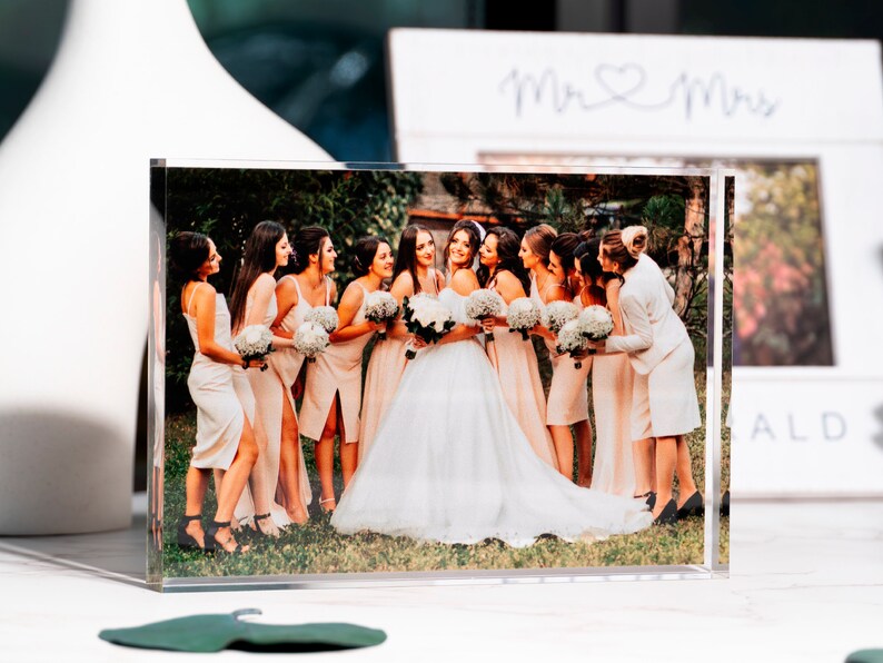 May include: A clear acrylic photo block displaying a wedding photo. The image features a bride in a white gown surrounded by bridesmaids in peach dresses, holding bouquets. A white vase and a framed sign with "Mr & Mrs" are in the background.
