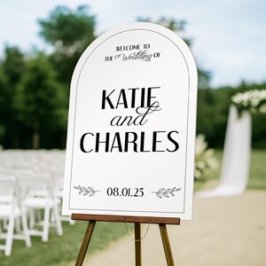 May include: A white wedding welcome sign on a wooden easel. The sign has an arched top and features the words "Welcome to The Wedding of KATIE and CHARLES" in black script. The date "08.01.25" is displayed below with leaf accents.