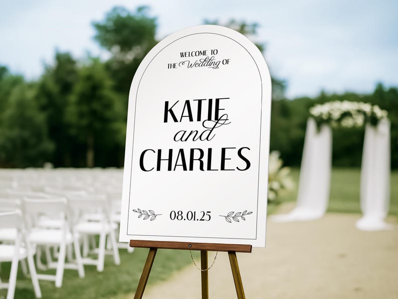 May include: A white wedding welcome sign on a wooden easel. The sign has an arched top and features the words "Welcome to The Wedding of KATIE and CHARLES" in black script. The date "08.01.25" is displayed below with leaf accents.