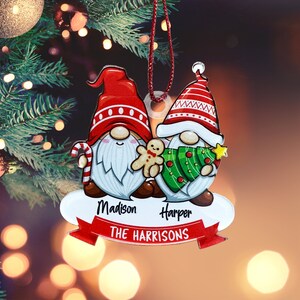 May include: Personalized Christmas ornament featuring two gnomes, one holding a gingerbread man and the other holding a Christmas tree. The ornament is decorated with red, green, and white colors and has the names "Madison" and "Harper" along with the text "THE HARRISONS".