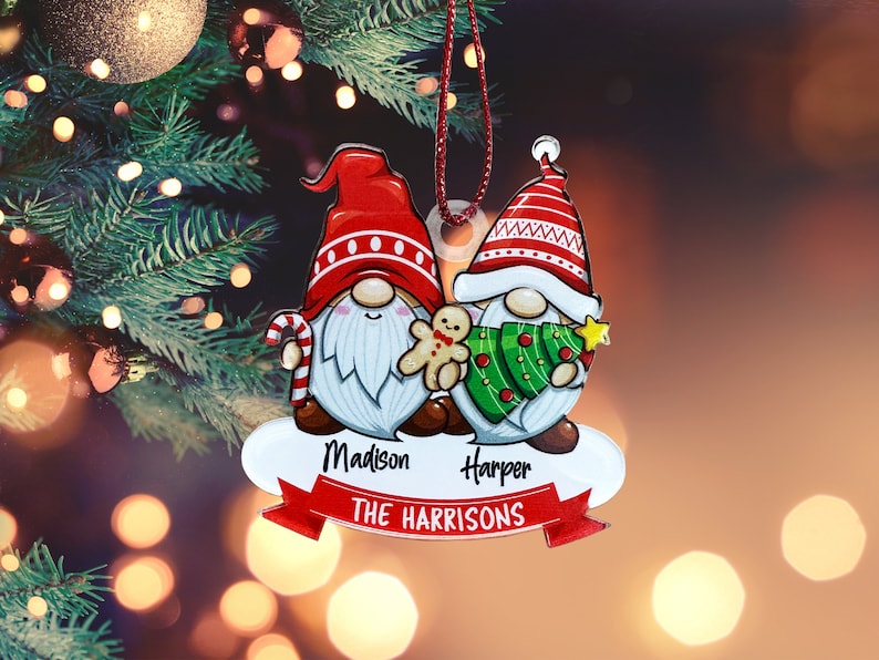 May include: Personalized Christmas ornament featuring two gnomes, one holding a gingerbread man and the other holding a Christmas tree. The ornament is decorated with red, green, and white colors and has the names "Madison" and "Harper" along with the text "THE HARRISONS".
