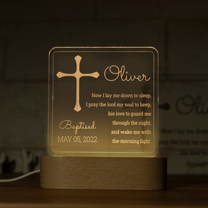 May include: A personalized baptism night light with a cross design and the name "Oliver". The light includes the date "MAY 05, 2022" and a prayer. It sits on a wooden base and emits a warm glow.