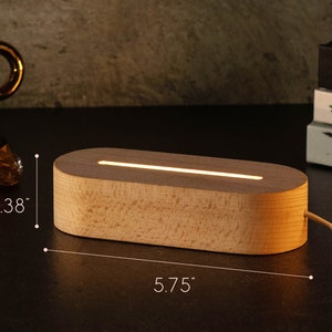 May include: A wooden, oval-shaped LED lamp with a warm, glowing light. The lamp is 5.75 inches long and 1.38 inches high. The light is emitting from a thin slit on the top of the lamp. A white cord extends from the back.