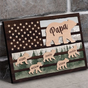 May include: Wooden wall art featuring a brown bear silhouette with the word "Papa" inside. The bear is standing in front of a stylized American flag with a forest scene below. Five smaller bear silhouettes are below the large bear, each with a name: Elijah, Willow, Noelle, Khloe, and Cecilia.