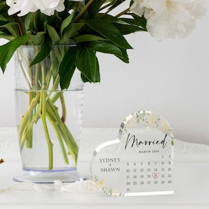 Puede incluir: Un calendario acr&iacute;lico transparente en forma de coraz&oacute;n con un dise&ntilde;o floral. El calendario es para marzo de 2024 y tiene los nombres de Sydney y Shawn escritos en &eacute;l. El 21 est&aacute; resaltado con un coraz&oacute;n rojo.