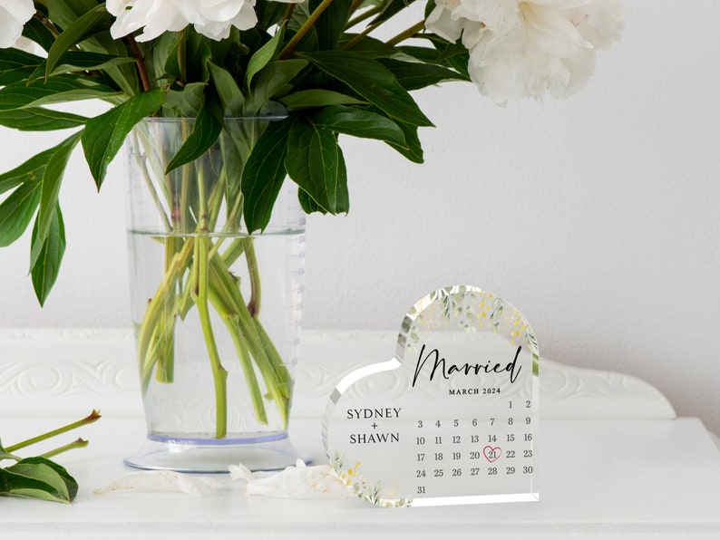 Puede incluir: Un calendario acr&iacute;lico transparente en forma de coraz&oacute;n con un dise&ntilde;o floral. El calendario es para marzo de 2024 y tiene los nombres de Sydney y Shawn escritos en &eacute;l. El 21 est&aacute; resaltado con un coraz&oacute;n rojo.