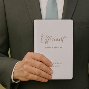 May include: A light gray notebook with the word "Officiant" in elegant script. Below, the name "RYAN JOHNSON" is printed. The bottom of the notebook includes the names "A & PAUL" and the date "03.23.2025". The notebook is held by a person wearing a dark green suit.