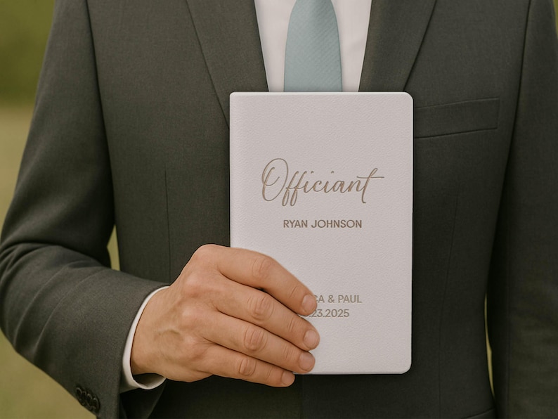 May include: A light gray notebook with the word "Officiant" in elegant script. Below, the name "RYAN JOHNSON" is printed. The bottom of the notebook includes the names "A & PAUL" and the date "03.23.2025". The notebook is held by a person wearing a dark green suit.