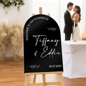 May include: A black wedding welcome sign on a wooden easel. The sign has white text that reads "Welcome to the Wedding of Tiffany & Eddie" and the date "06.27.2025". Decorative leaf accents are also included.