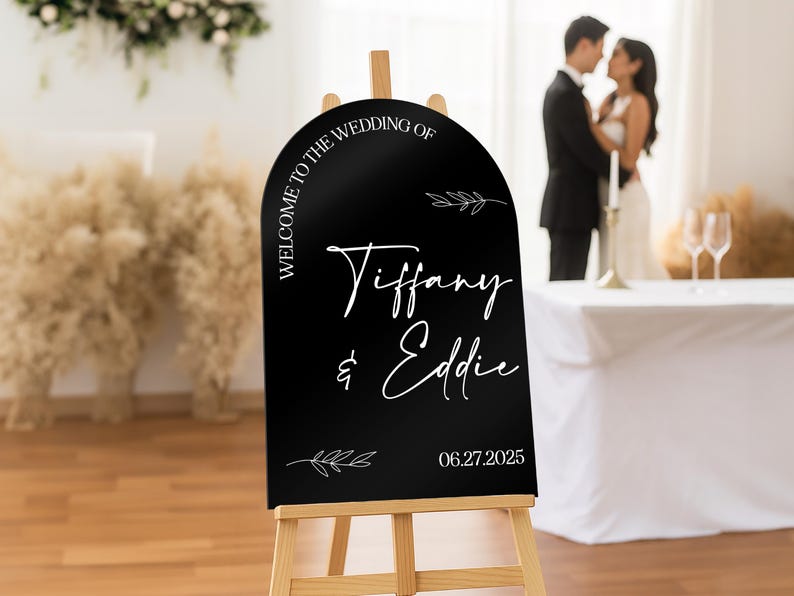 May include: A black wedding welcome sign on a wooden easel. The sign has white text that reads "Welcome to the Wedding of Tiffany & Eddie" and the date "06.27.2025". Decorative leaf accents are also included.